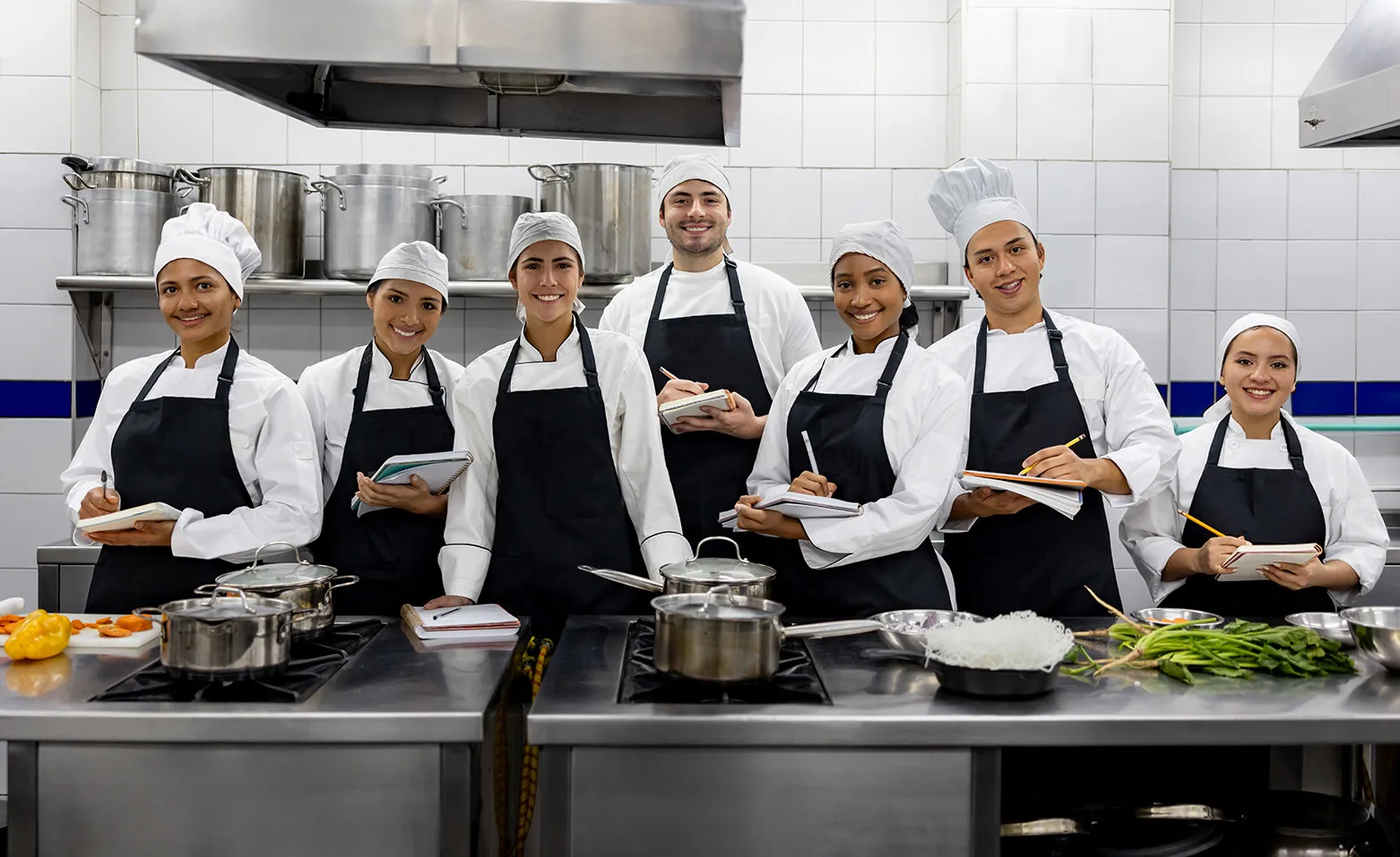 A group of smiling culinary workers in chef uniforms and aprons standing together in a professional kitchen, representing UNITE HERE members covered by the Adjustable Plan.