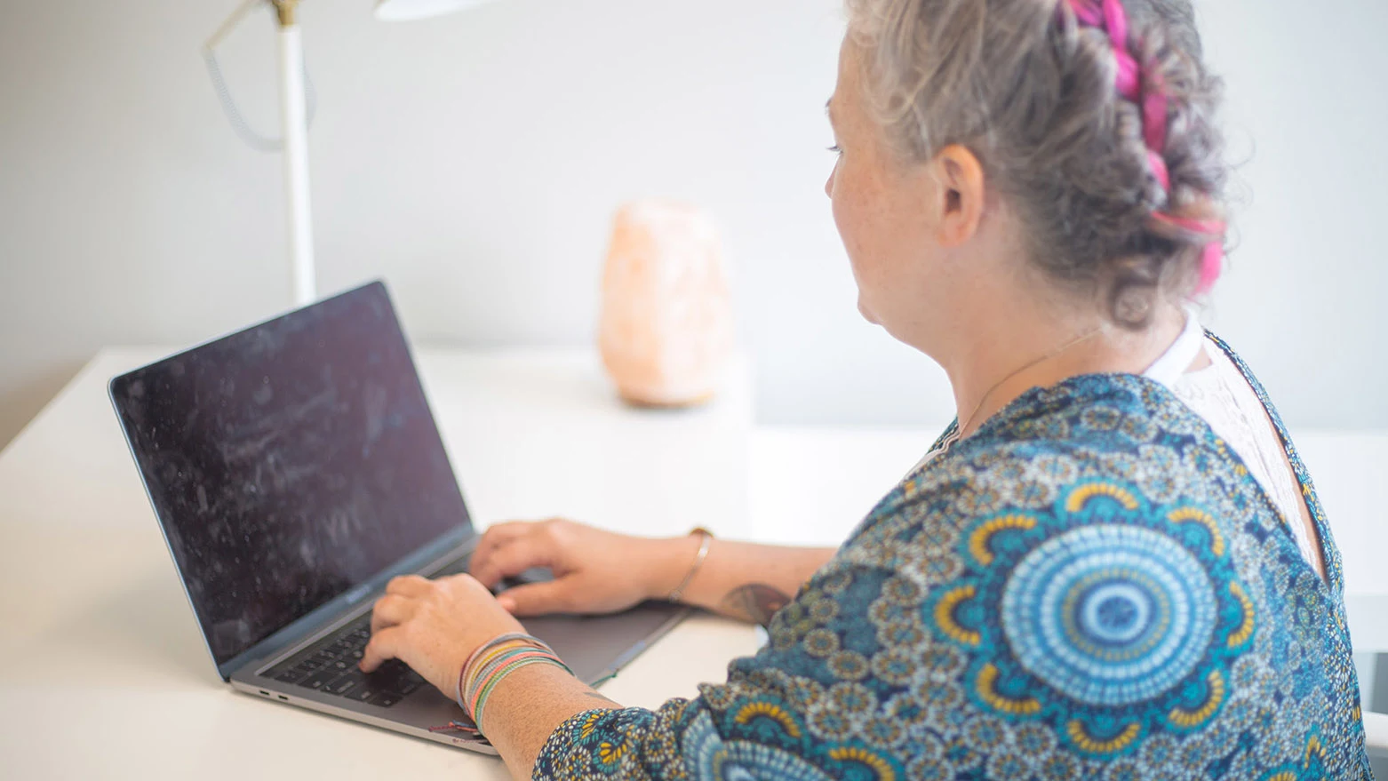 A person with colourful hair sitting at a desk and using a laptop to manage their pension account online.